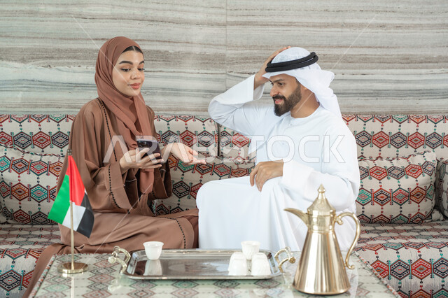 Expressions that remind of something, communication with family and friends, an Emirati Gulf Arab couple wearing traditional attire sitting in a popular ground council enjoying using their mobile phone, following news on social media, watching movies and series on their mobile.