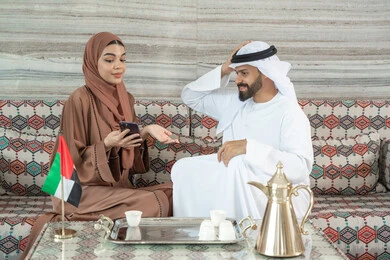 Expressions that remind of something, communication with family and friends, an Emirati Gulf Arab couple wearing traditional attire sitting in a popular ground council enjoying using their mobile phone, following news on social media, watching movies and series on their mobile.