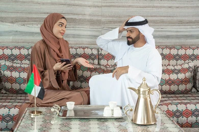 التواصل مع الأهل والأصدقاء، زوجان عربيان خليجيان اماراتيان يرتديان الزي التقليدي يجلسان فى مجلس ارضي شعبي يستمتعان باستخدام الهاتف المحمول، متابعة أخبار مواقع التواصل الاجتماعي، مشاهدة الافلام والمسلسلات عبر الجوال، تعابير تذكر شيء ما