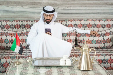 قضاء الوقت في تصفح السوشيال ميديا، انجاز المهام عن بعد، دمج التكنولوجيا في الحياة اليومية، رجل عربي خليجي اماراتي يرتدي الكندورة و الغتره يجلس فى مجلس ارضي شعبي يحمل بيده هاتف محمول ، الدردشة مع الأهل والأصدقاء عبر الانترنت