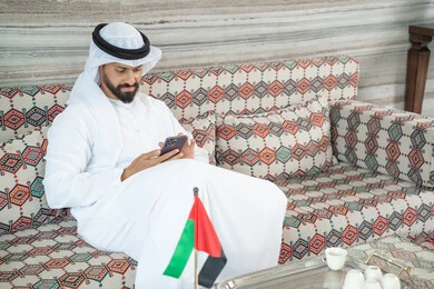 متابعة المنشورات على مواقع التواصل، انجاز المهام عن بعد، دمج التكنولوجيا في الحياة اليومية، رجل عربي خليجي اماراتي يرتدي الكندورة و الغتره يجلس فى مجلس ارضي شعبي يحمل بيده هاتف محمول ، الدردشة مع الأهل والأصدقاء عبر الانترنت