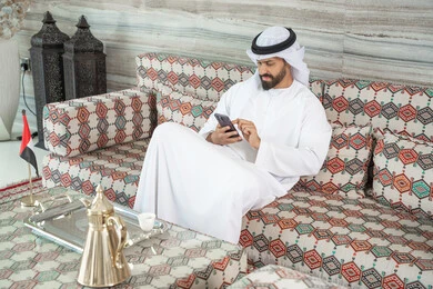 Browsing social media applications on mobile, completing tasks remotely, integrating technology into daily life, an Arab Gulf Emirati man wearing a kandura and ghutrah sitting in a traditional floor seating area holding a mobile phone, chatting with family and friends online. Browsing social media applications on mobile, completing tasks remotely, integrating technology into daily life, an Arab Gulf Emirati man wearing a kandura and ghutrah sitting in a traditional floor seating area holding a mobile phone, chatting with family and friends online.
