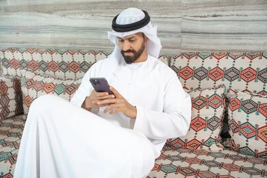 الدردشة مع الأهل والأصدقاء عبر الانترنت، انجاز المهام عن بعد، دمج التكنولوجيا في الحياة اليومية،رجل عربي خليجي اماراتي يرتدي الكندورة و الغتره يجلس فى مجلس ارضي شعبي يحمل بيده هاتف محمول ، تصفح وسائل التواصل الاجتماعي
