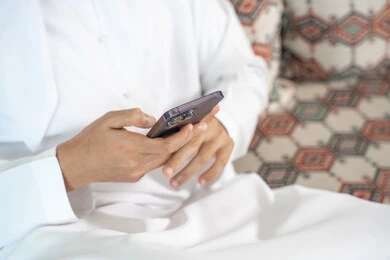 تصفح وسائل التواصل الاجتماعي، دمج التكنولوجيا في الحياة اليومية، انجاز المهام عن بعد، صورة مقربة ليد رجل عربي خليجي اماراتي يرتدي الكندورة  يحمل بيده هاتف محمول ، الدردشة مع الأهل والأصدقاء عبر الانترنت