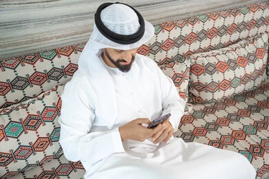 دمج التكنولوجيا في الحياة اليومية، انجاز المهام عن بعد، رجل عربي خليجي اماراتي يرتدي الكندورة و الغتره يجلس فى مجلس ارضي شعبي يحمل بيده هاتف محمول ، تصفح وسائل التواصل الاجتماعي ، الدردشة مع الأهل والأصدقاء عبر الانترنت