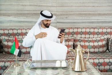 Communicating with friends through mobile applications, browsing social media sites, using advanced modern technology in professional life, consuming and drinking hot beverages, an Arab Gulf Emirati man wearing a kandura and ghutrah sitting in a traditional ground council holding a cup of coffee in his hands and using a mobile phone.