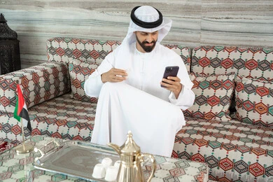 تصفح مواقع السوشال ميديا، استخدام التقنية الحديثه المتطورة في الحياة العملية، تناول وشرب المشروبات الساخنة، رجل عربي خليجي اماراتي يرتدي الكندورة و الغتره يجلس فى مجلس ارضي شعبي يمسك بيديه كوب قهوه و يقوم باستخدام الهاتف المحمول