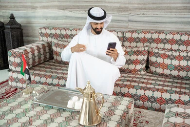 The use of advanced modern technology in practical life, consuming and drinking hot beverages, an Arab Gulf Emirati man wearing a kandura and ghutrah sitting in a traditional ground council holding a cup of coffee in his hands and using a mobile phone, browsing social media sites. The use of advanced modern technology in practical life, consuming and drinking hot beverages, an Arab Gulf Emirati man wearing a kandura and ghutrah sitting in a traditional ground council holding a cup of coffee in his hands and using a mobile phone, browsing social media sites.