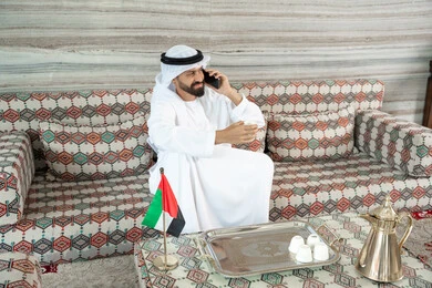 استخدام الأجهزة الحديثة والمتطورة، التواصل مع الأهل والأصدقاء, رجل عربي خليجي اماراتي يرتدي الكندورة و الغتره يجلس فى مجلس ارضي شعبي يقوم إجراء مكالمة صوتية عبر الهاتف المحمول, تعابير السعادة و السرور
