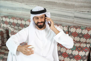 التواصل مع الأهل والأصدقاء, رجل عربي خليجي اماراتي يرتدي الكندورة و الغتره يجلس فى مجلس ارضي شعبي يقوم إجراء مكالمة صوتية عبر الهاتف المحمول, استخدام الأجهزة الحديثة والمتطورة, تعابير السعادة و السرور