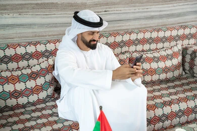 Completing tasks remotely, integrating technology into daily life, an Arab Gulf Emirati man wearing a kandura and ghutrah sitting in a traditional floor seating area holding a mobile phone, browsing social media, chatting with family and friends online. Completing tasks remotely, integrating technology into daily life, an Arab Gulf Emirati man wearing a kandura and ghutrah sitting in a traditional floor seating area holding a mobile phone, browsing social media, chatting with family and friends online.