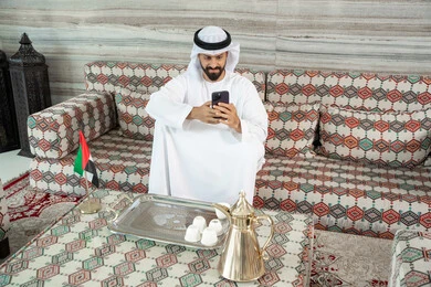 The use of mobile phones for communication with individuals, browsing the internet, and online shopping. An Arab Gulf Emirati man wearing a kandura and ghutrah sits in a traditional ground-level gathering holding a mobile phone in his hand. The use of mobile phones for communication with individuals, browsing the internet, and online shopping. An Arab Gulf Emirati man wearing a kandura and ghutrah sits in a traditional ground-level gathering holding a mobile phone in his hand.