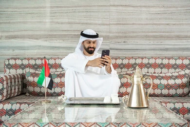 Browsing the internet and online shopping, using a mobile phone to communicate with individuals, an Arab Gulf Emirati man wearing a kandura and ghutrah sitting in a traditional ground council holding a mobile phone in his hand.