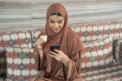 An Arab Gulf Emirati woman wearing an abaya and hijab sits in a traditional ground seating area holding a mobile phone and a cup of coffee, enjoying her favorite drink, with expressions of happiness and joy, communicating and exchanging conversations with family and friends, using a modern and advanced technological device.