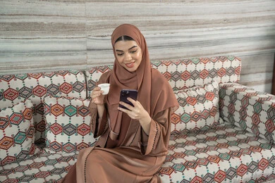الاستمتاع بشرب المشروب المفضل، تعابير السعادة و السرور، التواصل وتبادل الأحاديث مع الأهل والأصدقاء، استخدام جهاز تقني حديث ومتطور، امراه عربيه خليجيه اماراتيه ترتدي العباءة و الحجاب تجلس فى مجلس شعبي ارضي تمسك بيديها الهاتف المحمول وكوب من القهوة