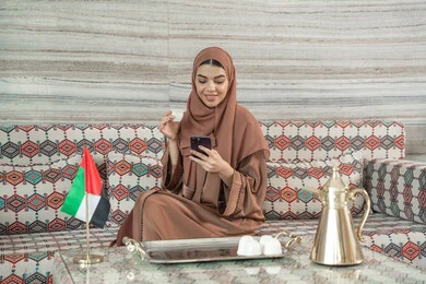 Expressions of happiness and joy, communication and exchanging conversations with family and friends, sipping a favorite drink, using a modern and advanced technological device, an Emirati Gulf Arab woman wearing an abaya and hijab sitting in a traditional ground seating area holding a mobile phone and a cup of coffee.