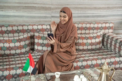 Communicating with family and friends online, using advanced modern technology via mobile, raising a hand in gestures of peace and greeting, expressions of happiness and joy, a young Arab Gulf Emirati woman wearing a hijab and abaya sitting in a traditional floor seating area making video calls online.