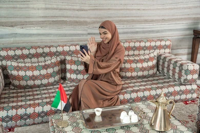 التحدث مع الأهل و الأصدقاء عن بعد، استخدام التقنية الحديثه المتطورة عبر الجوال، رفع كف الايد بايماءات السلام و التحيه، تعابير السعادة و السرور، شابة عربية خليجية اماراتية ترتدي الحجاب والعباءة تجلس فى مجلس ارضي شعبي تقوم باجراء مكالمات فيديو عبر الإنترنت