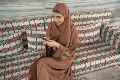 متابعة أخبار وسائل التواصل الاجتماعي باستخدام الجوال، إمرأة عربية خليجية اماراتية مبتسمة ترتدي حجاب وعباءة تجلس في مجلس ارضي شعبي تمسك بيديها الهاتف المحمول، تعابير وإيماءات الفرح والسرور، الدردشة والتواصل مع التواصل مع الأصدقاء أونلاين