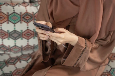 Using modern technical devices, chatting with family and friends, a close-up shot of the hand of an Arab Gulf Emirati woman wearing an abaya holding a mobile phone browsing social media.