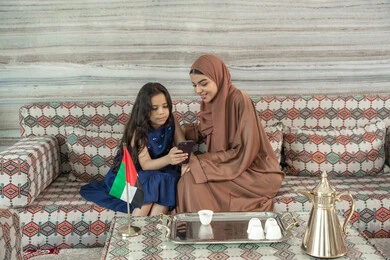 Watching favorite TV shows online, unique and special sessions, spending enjoyable times outdoors with children, an Emirati Gulf Arab woman wearing an abaya and hijab sitting in a traditional ground seating area with her daughter, both looking at a mobile phone, expressions of happiness and joy.