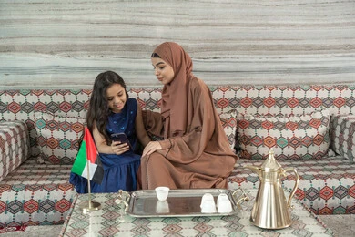 مشاهدة البرامج الترفيهية عبر المنصات الإلكترونية، جلسات فريدة و مميزة، قضاء اوقات ممتعة في الخارج مع الابناء, امراه عربيه خليجيه اماراتية ترتدي العباءة و الحجاب تجلس فى مجلس شعبي ارضي برفقة  ابنتها يقومون بالنظر الى الهاتف المحمول, تعابير السعاده و السرور