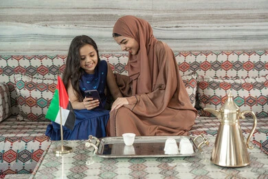 الاستمتاع بمشاهدة الافلام المفضلة أونلاين، جلسات فريدة و مميزة، قضاء اوقات ممتعة في الخارج مع الابناء, امراه عربيه خليجيه اماراتية ترتدي العباءة و الحجاب تجلس فى مجلس شعبي ارضي برفقة  ابنتها يقومون بالنظر الى الهاتف المحمول, تعابير السعاده و السرور
