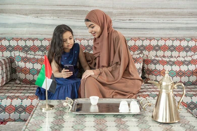 Watching series and programs online, unique and special sessions, spending enjoyable times outdoors with children, an Arab Gulf Emirati woman wearing an abaya and hijab sitting in a traditional ground seating area with her daughter, both looking at a mobile phone, expressions of happiness and joy.