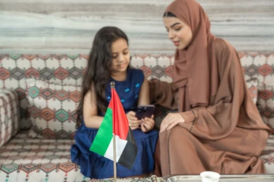 Watching favorite programs using smart devices, unique and special sessions, spending enjoyable times outdoors with children, an Arab Gulf Emirati woman wearing an abaya and hijab sitting in a traditional ground seating area with her daughter, both looking at a mobile phone, expressions of happiness and joy.