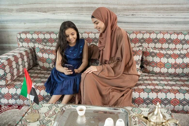 الاستمتاع بمشاهدة البرامج عبر المنصات الرقمية، جلسات فريدة و مميزة، قضاء اوقات ممتعة في الخارج مع الابناء, امراه عربيه خليجيه اماراتية ترتدي العباءة و الحجاب تجلس فى مجلس شعبي ارضي برفقة  ابنتها يقومون بالنظر الى الهاتف المحمول, تعابير السعاده و السرور