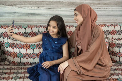 Capturing a family selfie to preserve memories, the joy of children spending enjoyable time with their family, an Emirati Gulf Arab girl wearing a dress sitting in a traditional floor seating area documenting fun moments with her mother, gestures of happiness and joy.