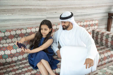التقاط سيلفي عائلي تذكاري عبر الهاتف المحمول، فرحة الأبناء بقضاء وقت ممتع مع الأهل, بنت عربيه خليجيه اماراتية ترتدي الفستان تجلس فى مجلس ارضي شعبي تقوم بتوثيق اللحظات الممتعة مع والدها ، ايماءات السعادة و السرور