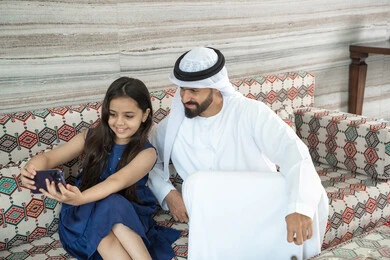 The joy of children spending quality time with family, taking a memorable family selfie with a mobile phone, an Emirati Gulf Arab girl wearing a dress sitting in a traditional floor seating area documenting fun moments with her father, gestures of happiness and joy.