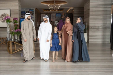 استخدام اليد للدلالة على شيء، الاستمتاع باللحظات الأسرية السعيدة، الاستعداد لقضاء رحلة سياحية ممتعه مع العائلة، تبادل الاحاديث بتعابير السرور و السعادة,عائلة عربيه خليجيه اماراتية ترتدي الزي التقليدي يسيرون داخل مدخل الاستقبال فى الفندق