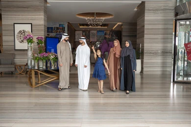 Pointing the signal towards something specific, enjoying happy family moments, preparing to spend an enjoyable tourist trip with the family, exchanging conversations with expressions of joy and happiness, an Emirati Gulf Arab family wearing traditional attire walking inside the hotel reception entrance.