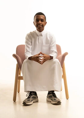 The concept of elegance, masculinity, and attention to appearance; a portrait of a young Arab Gulf Saudi teenager wearing traditional attire, smiling while sitting on a comfortable chair, looking at the camera and pointing at something, with expressions of joy and happiness, against a white background.
