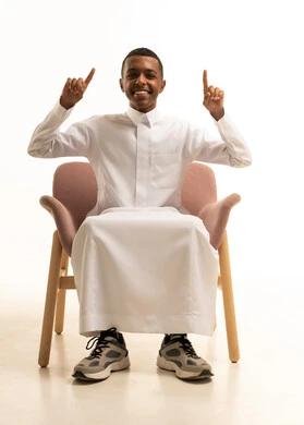 اشارات وحركات يدين تدل على هذا المكان (هنا)، تحديد موقع معين بتعابير البشاشة، بورتريه لشاب مراهق عربي خليجي سعودى مبتسم يرتدي الثوب التقليدى يجلس على كرسي مريح يرفع اصابع السبابة ويشير الى الاعلى، خلفية بيضاء