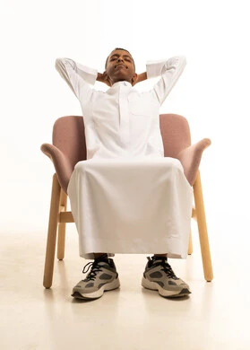 A close-up portrait of a young Arab Gulf Saudi teenager wearing traditional attire, sitting on a comfortable chair with his hands behind his head, embodying relaxation and enjoyment of the calm atmosphere, feeling tired and in need of sleep, with a white background.