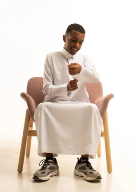 ترتيب اكمام الثوب، مفهوم الاناقة والرجولة والاهتمام بالمظهر الخارجي، بورتريه لشاب مراهق عربي خليجي سعودى مبتسم يرتدي الثوب التقليدى يجلس على كرسي مريح يقوم بحركات يدين مختلفة بتعابير السرور، خلفية بيضاء