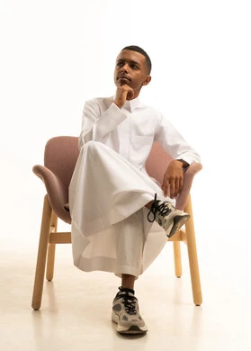 Gestures of confusion and anxiety, a portrait of a young Arab Gulf Saudi teenager wearing traditional attire sitting on a comfortable chair with his hand on his cheek, trying to find a solution to a problem, pondering, focusing, and planning for the future, expressions of thought and contemplation, mind wandering somewhere, white background.
