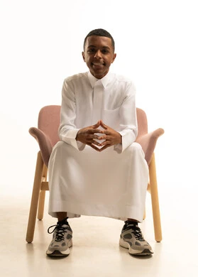 تعابير السرور و الفرح، مفهوم الاناقة والرجولة والاهتمام بالمظهرالخارجي، بورتريه لشاب مراهق عربي خليجي سعودى يرتدي الثوب التقليدى مبتسم يجلس على كرسي مريح ينظر الى الكاميرا يشير الى شىء ما، خلفية بيضا