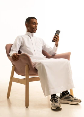 بورتريه لشاب عربي خليجي سعودي يرتدي الثوب التقليدي يجلس على كرسي مريح يمسك الهاتف المحمول بتعابير السعادة، استخدام الأجهزة والوسائل التقنية الحديثة، إرسال الرسائل النصية والصوتية عبر الجوال، مفهوم الجلوس والراحة،, خلفية بيضاء