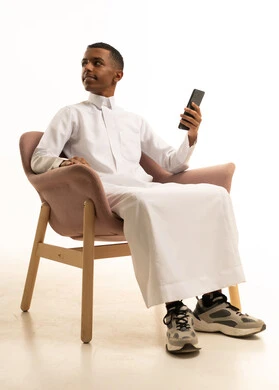 استخدام الأجهزة والوسائل التقنية الحديثة، إرسال الرسائل النصية والصوتية عبر الجوال، مفهوم الجلوس والراحة، بورتريه لشاب عربي خليجي سعودي يرتدي الثوب التقليدي يجلس على كرسي مريح يمسك الهاتف المحمول بتعابير السعادة, خلفية بيضاء