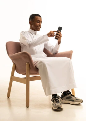 إرسال الرسائل النصية والصوتية عبر الجوال، مفهوم الجلوس والراحة، استخدام الأجهزة والوسائل التقنية الحديثة، بورتريه لشاب عربي خليجي سعودي يرتدي الثوب التقليدي يجلس على كرسي مريح يمسك الهاتف المحمول بتعابير السعادة, خلفية بيضاء