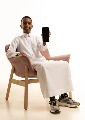 A display of a blank black screen, expressions and gestures of happiness and joy, a portrait of a smiling young Arab Gulf Saudi teenager wearing traditional attire, holding a mobile phone in his hand, sitting on a comfortable chair, using a modern advanced technological device, chroma, white background.