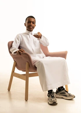 A portrait of a smiling young Saudi Gulf Arab sitting on a chair holding a TV remote control, engaging in recreational activities, enjoying fun times, watching movies, series, and entertainment programs on TV, with a white background.
