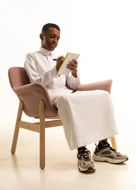 Expressions of diligence and excellence, reviewing lessons for studying and school tests, developing educational curricula in Saudi Arabia, a portrait of a young Saudi Arab teenager wearing traditional attire sitting on a comfortable chair writing in a textbook, gestures of focus and thought, white background.