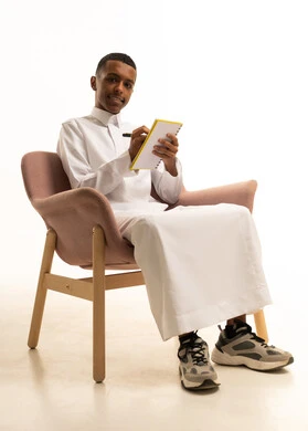 مراجعة الدروس للمذاكرة والاختبارات المدرسية، تطوير المناهج التعليمية في السعودية، تعابير الاجتهاد والتفوق, بورتريه لشاب مراهق عربي سعودى مبتسم يرتدي الثوب التقليدى يجلس على كرسي مريح يقوم بالكتابة على الكتاب المدرسي، خلفية بيضاء