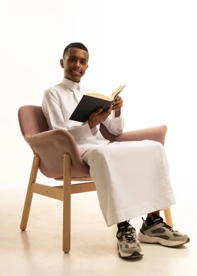 ايماءات التركيز و التفكير، تطوير المناهج التعليمية في السعودية، مراجعة الدروس للمذاكرة والاختبارات المدرسية، تعابير الاجتهاد والتفوق, بورتريه لشاب مراهق عربي خليجي سعودى مبتسم يرتدي الثوب التقليدى يجلس على كرسي مريح يمسك بيده الكتاب المدرسي، خلفية بيضاء