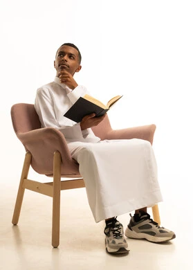 A portrait of a young Arab Gulf Saudi teenager wearing traditional attire sitting on a chair holding a school book in his hand, trying to remember something, the development of educational curricula in Saudi Arabia, gestures of diligence and excellence, reviewing lessons for studying, white background.